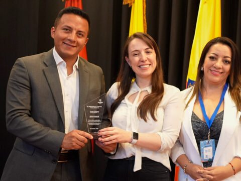 El secretario de gobierno de Sandoná, John Jairo Castillo, y la comisaria de familia, Diana Fernanda Delgado Pabón, reciben un reconocimiento por buenas prácticas de manos de una representante del Ministerio de Justicia y del Derecho, en un evento que destaca el trabajo de varias comisarías de familia en Colombia. Los tres posan sonrientes con el premio, con banderas de Colombia al fondo